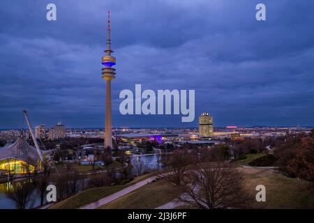 Vista dall'Olympiaberg con Torre Olimpica, illuminata in colori nazionali ucraini blu e giallo in occasione della guerra in Ucraina, BMW Welt e Allianz Arena, serata, capitale Monaco, Baviera, Germania, Europa Foto Stock