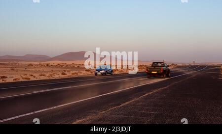 Spagna, Isole Canarie, Fuerteventura, zona delle dune di El Jable, strada accanto alla zona delle dune, sabbia soffiata sulla strada, auto sul lato della strada, pick-up furred guida in auto nella foto, azzurro cielo senza nuvole Foto Stock