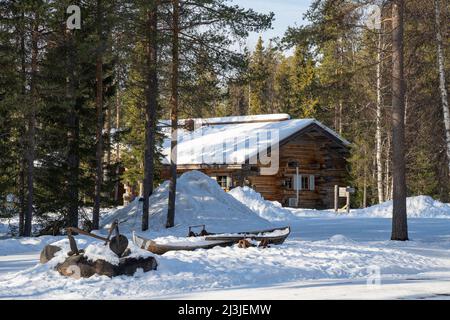 Rovaniemi, Finlandia - 17th marzo 2022: Una cabina di legno e una vecchia barca in una foresta di abeti innevati in una giornata invernale soleggiata. Foto Stock