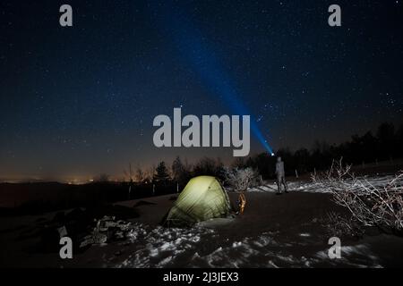 Uomo in piedi vicino alla tenda illuminata e puntando la torcia blu al cielo stellato in inverno Nebrodi Park, Sicilia Foto Stock