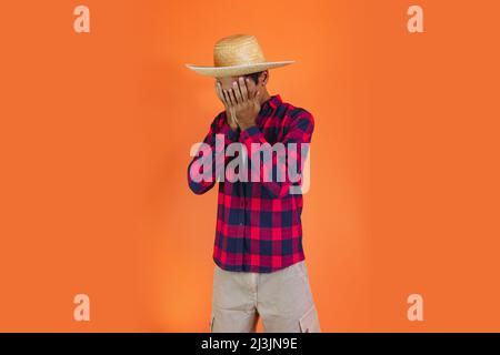 Black Man con Junina Party vestito isolato su sfondo arancione. Giovane uomo che indossa abiti tradizionali per Festa Junina - Festival brasiliano di giugno. Foto Stock