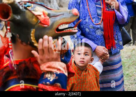 Un giovane ragazzo guarda in su come un uomo su una maschera per eseguire una danza Cham al Bhutanese Paro Tshechu Festival all'interno di Paro Dzong, un monastero e fortres... Foto Stock