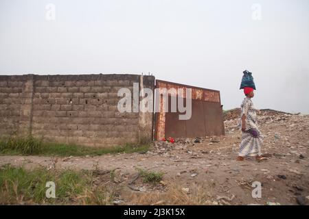 Una donna con la borsa caricata sulla sua testa cammina oltre una discarica di immondizia.; Kinshasa, Repubblica democratica del Congo. Foto Stock