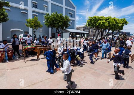 I bambini delle scuole in uniforme si fermano per ascoltare un gruppo musicale di percussioni lungo il lungomare di Città del Capo; Città del Capo, Sudafrica Foto Stock