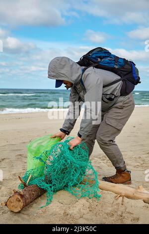 Il volontario maschile in tempo ventoso raccoglie rifiuti gettato da una tempesta su una spiaggia di sabbia. Il problema dell'inquinamento degli oceani e degli ambientani del mondo Foto Stock