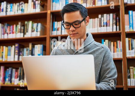 È uno studente di prima classe. Scatto di un giovane concentrato impegnato a lavorare sul suo laptop e studiare mentre si siede all'interno di una biblioteca. Foto Stock