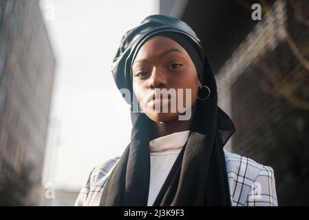 Vista laterale basso angolo di sicurezza afro-americana musulmana donna vestito in elegante scacchi blazer con foulard Islamico e berretto in pelle guardando la macchina fotografica con fiducia Foto Stock