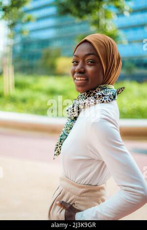 Affascinante femmina afro-americana musulmana in hijab tradizionale ed elegante sciarpa legata al collo in piedi sulla strada della città e fissando alla macchina fotografica Foto Stock