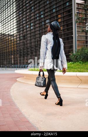 Back view full body of African American femmina in abito alla moda con foulard musulmano a piedi con borsa verso moderno edificio d'affari nel parco della città Foto Stock