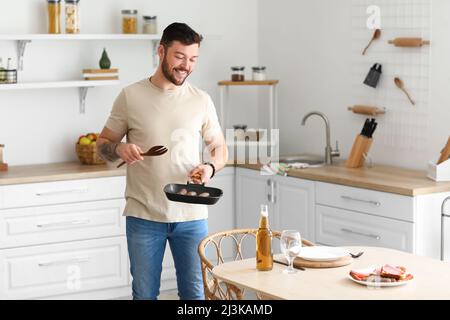 Giovane uomo portico con salsicce fritte vicino al tavolo in cucina Foto Stock