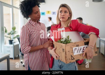 Una giovane impiegato femminile in un'atmosfera triste in ufficio sta confortando la sua giovane collega che viene licenziata. Dipendenti, lavoro, ufficio Foto Stock