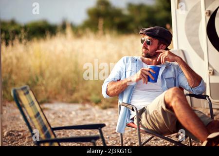 Giovane uomo seduto di fronte a camper e drink.Travel e concetto di amicizia celebrazione Foto Stock