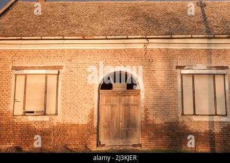 Foto di una fattoria abbandonata in Vojvodina, in Serbia, con la facciata della sua casa principale gravemente danneggiata e decadente. Foto Stock