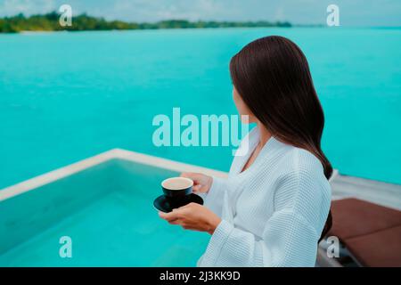 Donna che fa colazione in camera d'hotel di lusso bevendo una tazza di caffè con vista dell'oceano turchese e della piscina a sfioro sull'acqua. Vacanza in paradiso Foto Stock