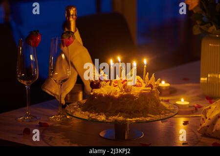 Tavolo con candele illuminate su una torta e bottiglia di champagne con bicchieri di champagne con fragola in un ambiente fioca; Germania Foto Stock