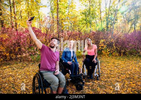 Giovane paraplegico uomo e donne in carrozzina prendendo un autoritratto con uno smartphone in un parco in una bella giornata di autunno Foto Stock