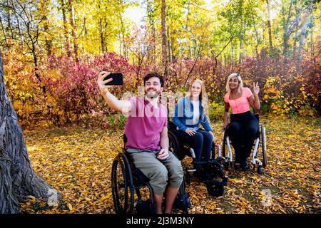 Giovane paraplegico uomo e donne in carrozzina prendendo un autoritratto con uno smartphone in un parco in una bella giornata di autunno Foto Stock