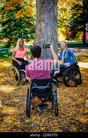 Giovane paraplegico uomo e donne in sedia a rotelle che scattano foto con uno smartphone in un parco in una bella giornata autunnale; Edmonton, Alberta, Canada Foto Stock