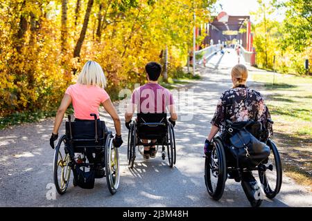 Tre giovani amici paraplegici trascorrono del tempo insieme percorrendo un sentiero in carrozzina in un parco cittadino in una splendida giornata autunnale Foto Stock