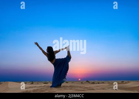 Donna con gonna lunga illuminata dal tramonto sulle sabbie desertiche, in piedi in una posizione di ballo yoga; Abu Dhabi, Emirati Arabi Uniti Foto Stock