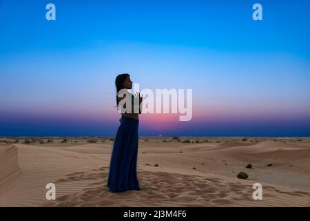 La donna si erge sulle sabbie del deserto contro un tramonto incandescente nella posa di preghiera yoga; Abu Dhabi, Emirati Arabi Uniti Foto Stock