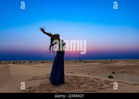 Donna con gonna lunga illuminata dal tramonto sulle sabbie del deserto, in piedi in una posa yoga curva posteriore; Abu Dhabi, Emirati Arabi Uniti Foto Stock