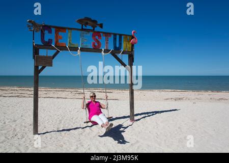 Donna su un altalena con segno Celestun su una spiaggia di sabbia bianca sulla costa del Golfo del Messico; Celestun, Yucatan Stato, Messico Foto Stock
