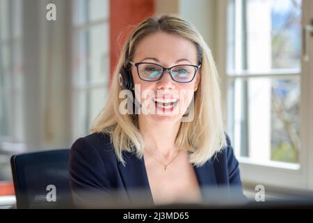 Donna d'affari sorridente felice che indossa occhiali e una cuffia che si siede in ufficio ridendo alla fotocamera con un'espressione divertente Foto Stock