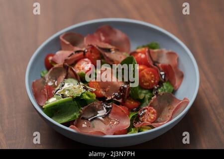 Insalata con bresaola, pomodori ciliegini e lattuga romena in ciotola blu su tavola di noce, fuoco basso Foto Stock