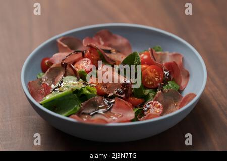 Insalata con bresaola, pomodori ciliegini e lattuga romena in ciotola blu su tavola di noce, fuoco basso Foto Stock