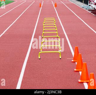 Undici ostacoli di plastica gialli alti sei pollici sistemati nella corsia quattro per lo sprinter della scuola superiore per correre sopra con coni arancioni nella distanza che contrassegna la pinna Foto Stock