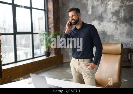 Sorridente uomo manager contemporaneo di successo in abbigliamento casual tenendo una telefonata. Felice ispirato caucasico maschio piccola impresa proprietario parlare sullo smartphone mentre in piedi in un moderno ufficio loft Foto Stock