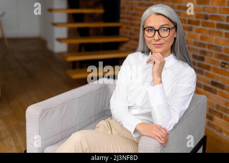 Incantevole donna spensierata di mezza età dai capelli grigi seduta sulla poltrona e rilassante. Ritratto di affascinante donna matura daydreaming a casa o in ufficio Foto Stock