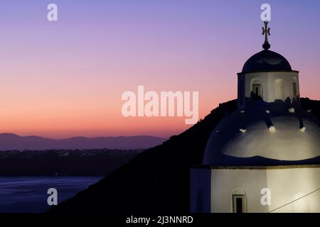 Vista di una bella chiesa ortodossa bianca e Oia Santorini sullo sfondo, mentre il sole tramonta in modo drammatico Foto Stock