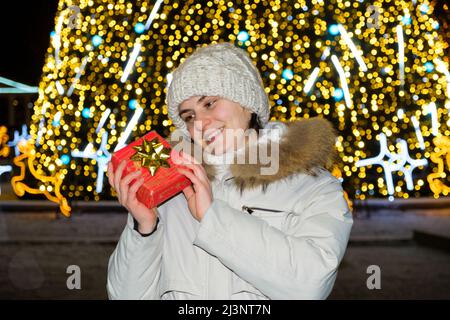 Una donna in abiti bianchi d'inverno si erge fuori da un albero di Natale, contiene una scatola regalo rossa e sorride. Foto Stock