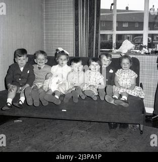 1962, storico, battesimo di famiglia, un gruppo di bambini piccoli seduti insieme su un divano in una linea per una foto, il bambino castigato che giace sul grembo di una bambina, Stockport, Manchester, Inghilterra, Regno Unito. Per l'occasione, domenica migliori abiti e sandali ai piedi. Foto Stock