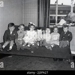 1962, storico, battesimo di famiglia, un gruppo di bambini piccoli seduti insieme su un divano in una linea per una foto, il bambino castigato che giace sul grembo di una bambina, Stockport, Manchester, Inghilterra, Regno Unito. Per i bambini, i migliori abiti e sandali. Foto Stock