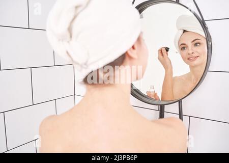 Trattamento pelle del viso della giovane donna. Massaggio sano con olio di Argan. Routine bagno mattina. Donna che applica il prodotto maschera siero. Liquido cosmetico contagocce terapia. Foto con specchio Foto Stock