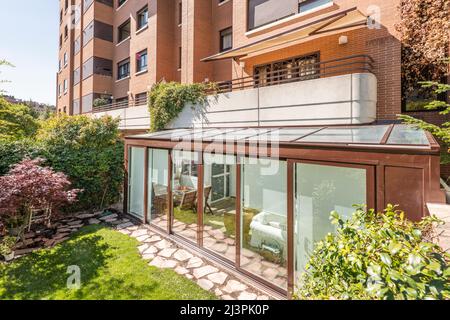 Terrazza marrone in alluminio e vetro coperta nella parte inferiore di un palazzo residenziale con porte scorrevoli, alberi decorativi e un sentiero acciottolato Foto Stock