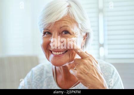 Ora è il momento di prendere cura della pelle. Ritratto di una donna anziana felice che applica crema di pelle al suo viso a casa. Foto Stock