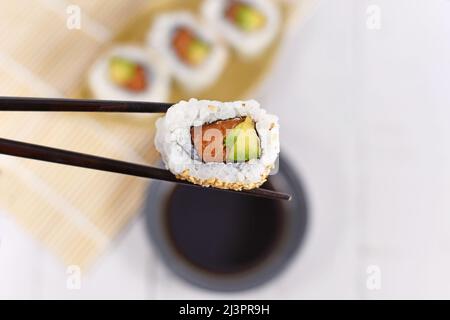 Sushi singolo all'interno con salmone, avocado e sesamo tenuti da bacchette Foto Stock