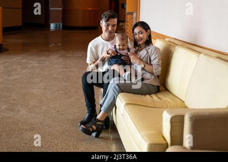 Malay Asian Malese Mussulmano giovane coppia con un bambino di un anno di età figlia seduta su una lunga sedia marrone chiaro divano, capelli lunghi, tutta la lunghezza o vita in su Foto Stock