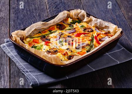 al forno frittata di verdure con uova sbattute, cipolla rossa, patate dolci, broccoli, peperoni rossi e funghi tagliati in una teglia da forno su tavola di legno scuro Foto Stock