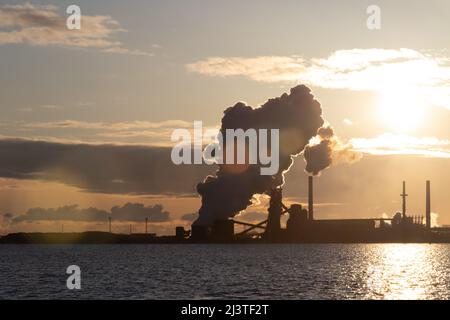 Gli edifici industriali e i fumetti sono visti dall'altra parte dell'acqua al mattino presto, mentre il sole splende. Foto Stock