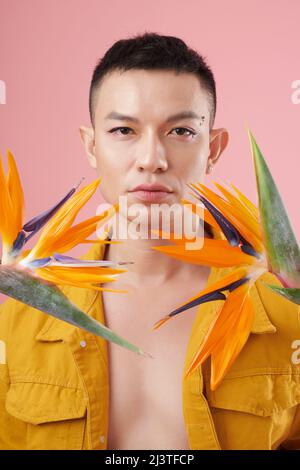 Ritratto di giovane uomo che indossa eyeliner e rhinestones trucco quando in piedi tra i fiori di strelitzia Foto Stock