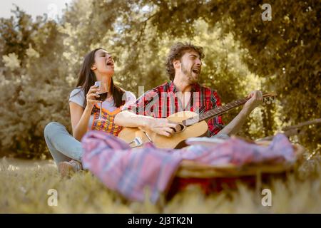 Coppia di giovani amici ubriachi bevendo vino rosso e suonando la chitarra e cantando all'aperto divertirsi insieme durante un picnic nel parco. Foto Stock