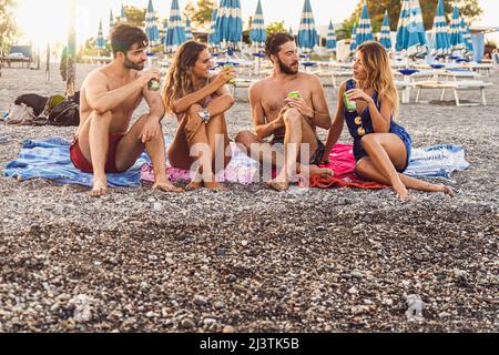Due coppie di giovani trascorrono felicemente del tempo su una spiaggia di ciottoli parlando insieme e bevendo birre in scatola - i millennial si riuniscono al fine settimana per andare Foto Stock