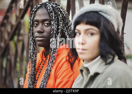 Multietnico diverse coppie di donne seduti all'aperto e guardando seriamente la macchina fotografica - Biracial girlfriends in piedi all'aperto accanto a un cancello - urbano Foto Stock