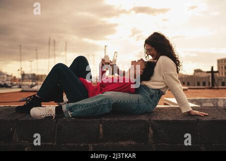 Coppia di amiche che si divertono seduti sulla parete del porto, bevendo birre e spensierate - gente di gen z godersi la vita Foto Stock