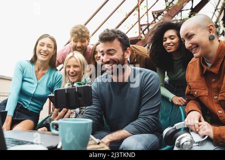 Gruppo di studenti universitari seduti intorno a uno smartphone su un piccolo treppiede in streaming online e ridendo insieme guardando le statistiche sul cellulare Foto Stock